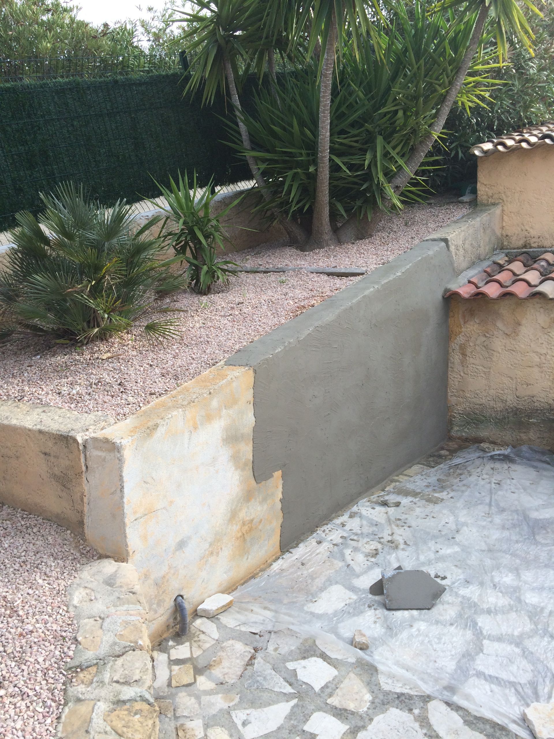 Mur extérieur en cours de rénovation avec une partie recouverte de plâtre gris et l'autre partie en béton brut. Jardin avec des plantes en arrière-plan.