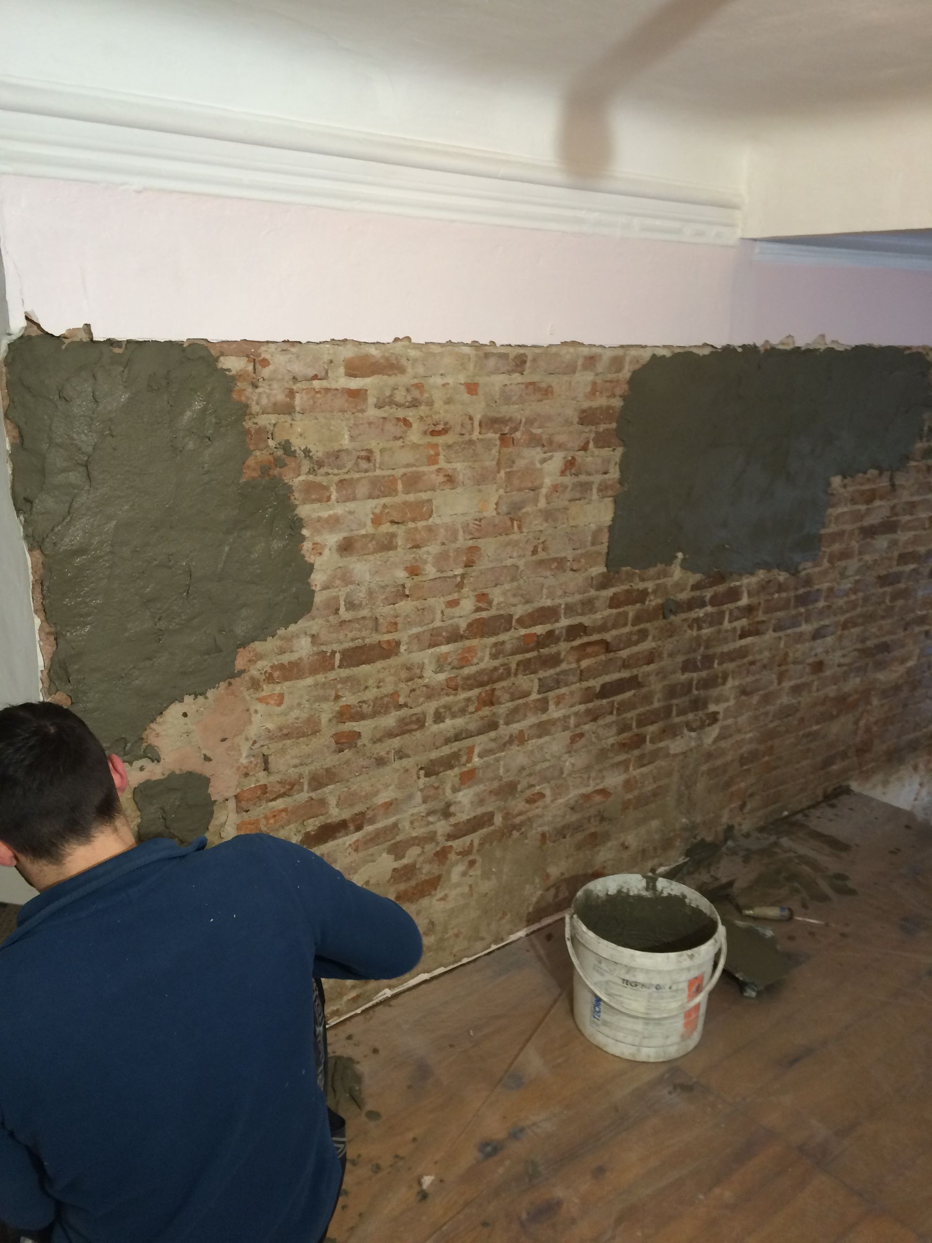 Un homme applique du mortier sur un mur en briques apparentes. Un seau de mortier est visible au sol.