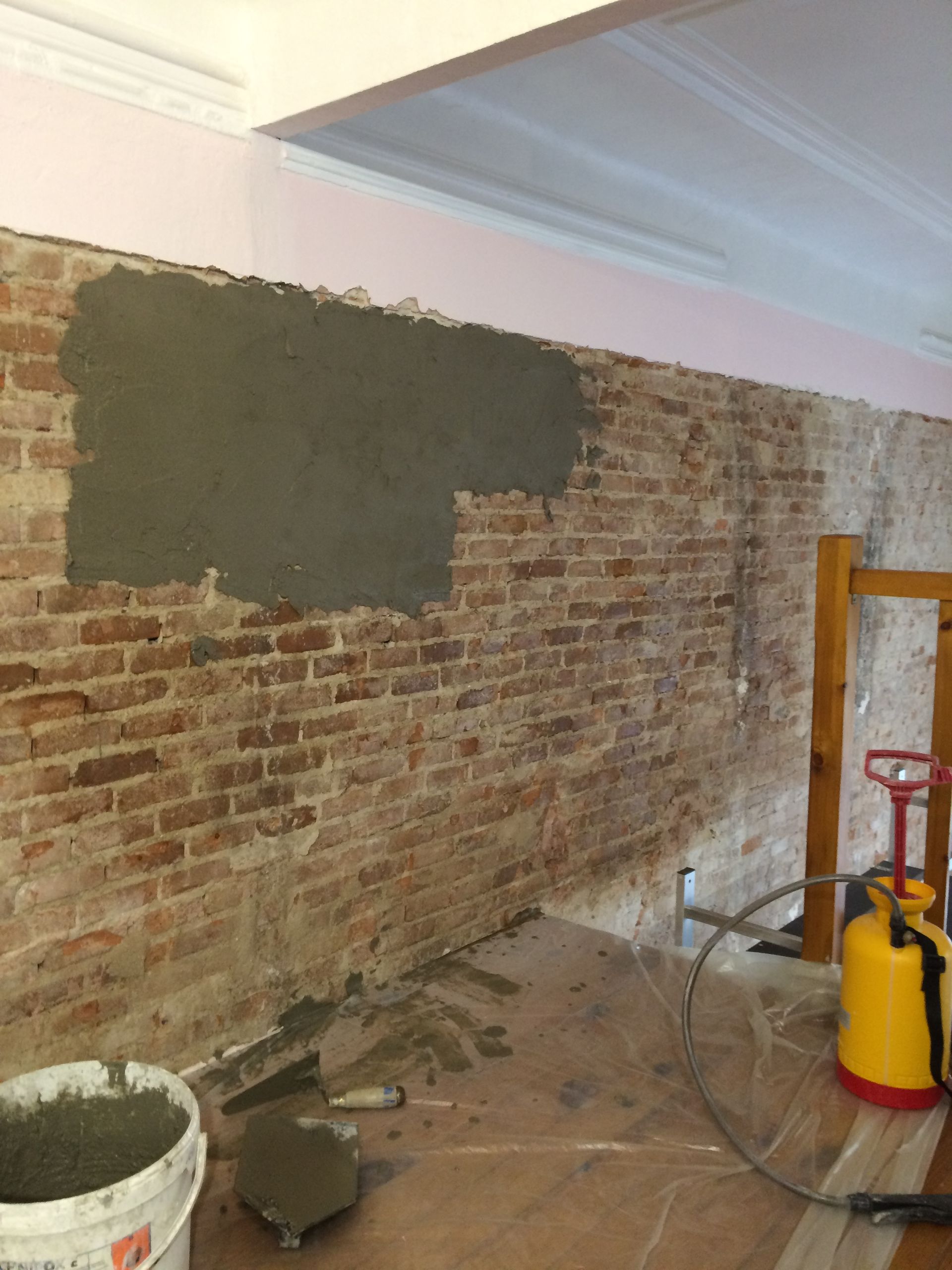 
Un mur en briques partiellement recouvert de mortier, avec des outils de construction au sol.
