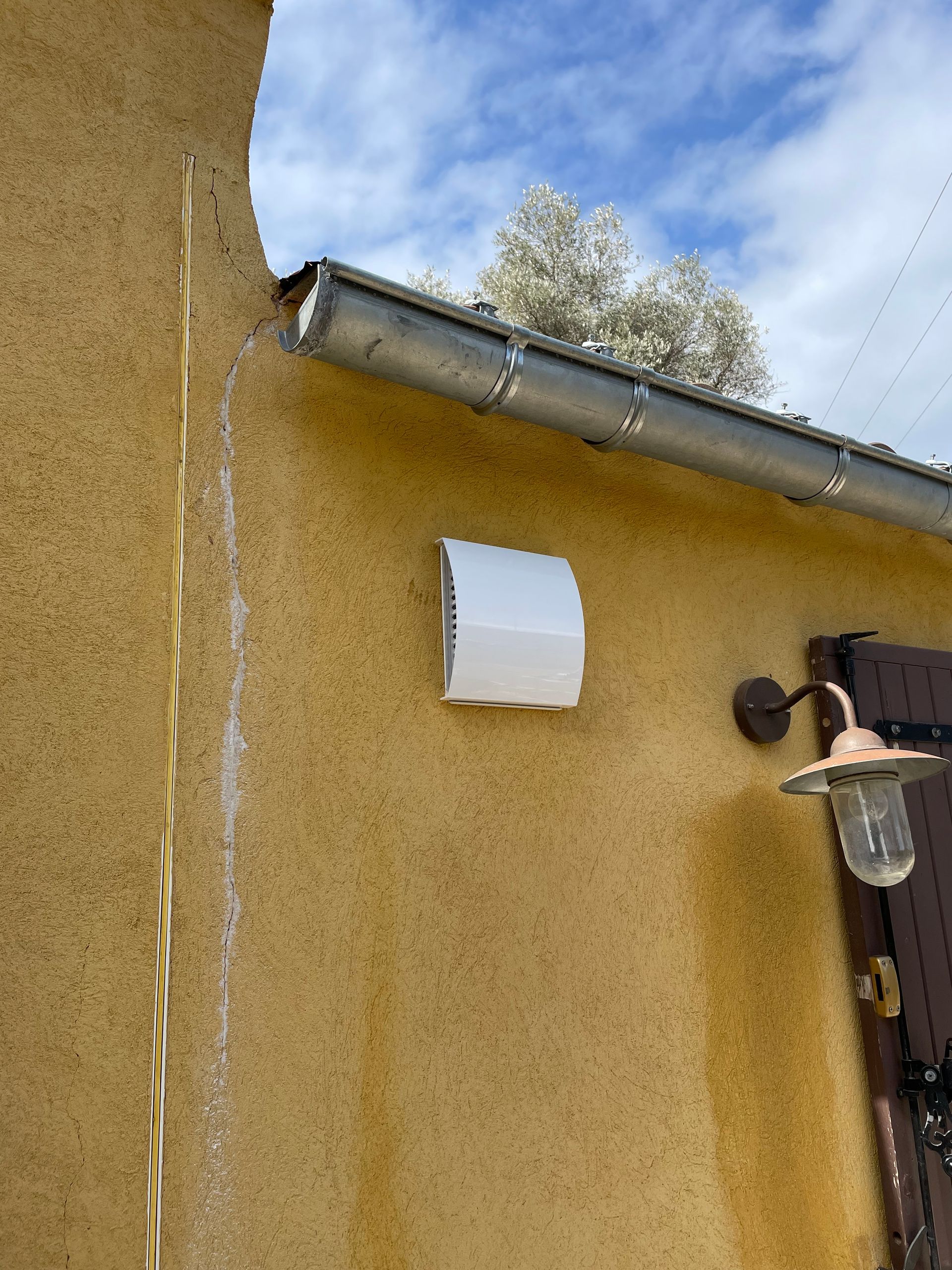Mur extérieur beige avec gouttière métallique et conduit d'aération blanc.