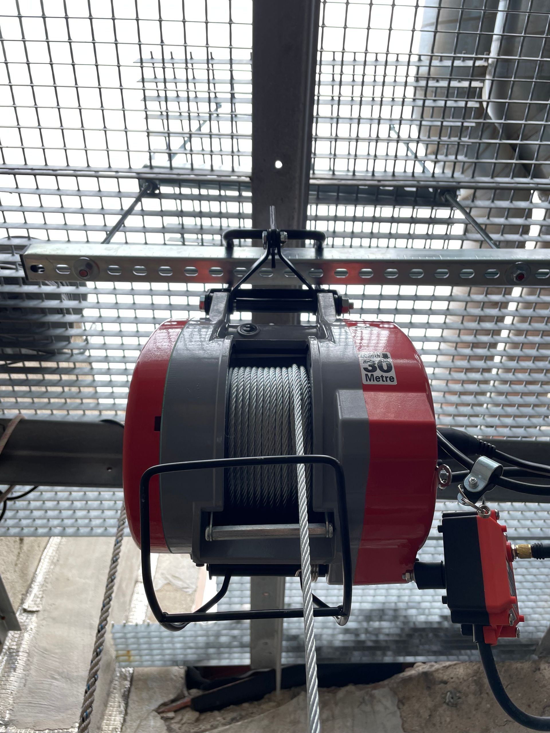 Vue d'un dispositif mécanique rouge et gris fixé à une structure métallique. Le dispositif semble être un treuil ou un système de levage