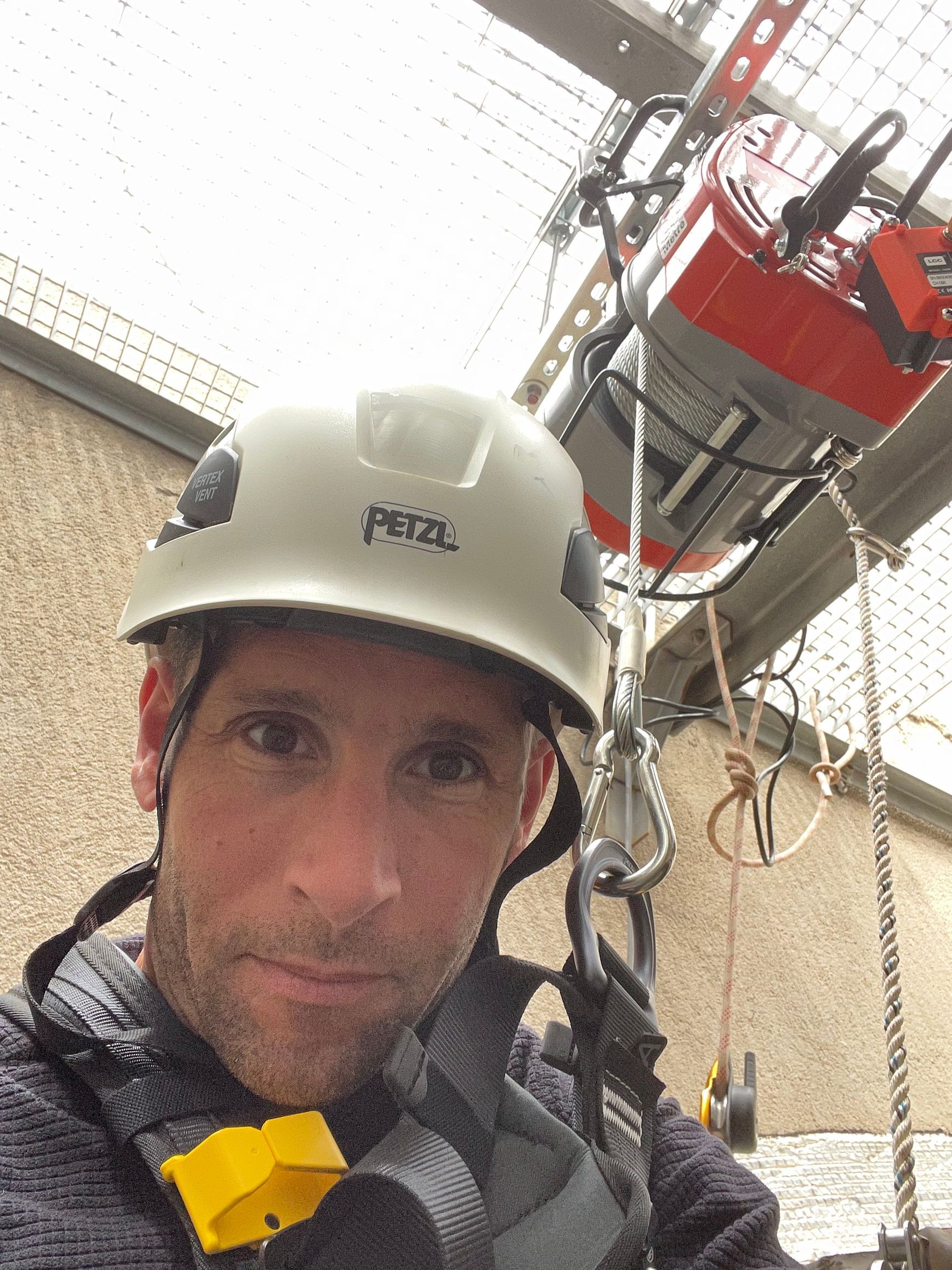 Selfie d'un ouvrier de Humidi T portant un casque de sécurité blanc et un harnais de sécurité. La personne est devant une structure métallique avec un treuil.