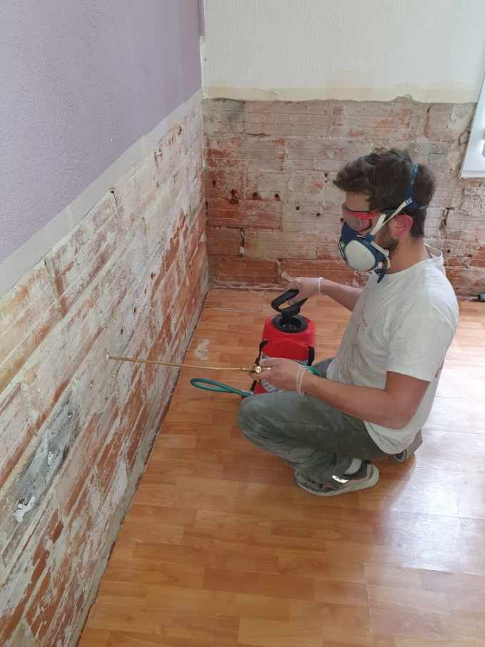 Un homme utilise un pulvérisateur pour appliquer un produit sur un mur en briques apparentes. Il porte un masque de protection et des lunettes de sécurité.