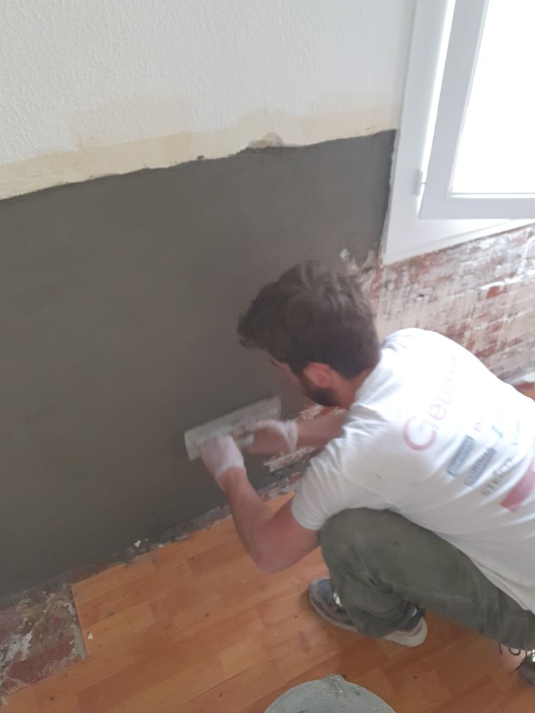 Un homme applique du plâtre sur un mur intérieur. Il est agenouillé et utilise une truelle pour lisser le plâtre.