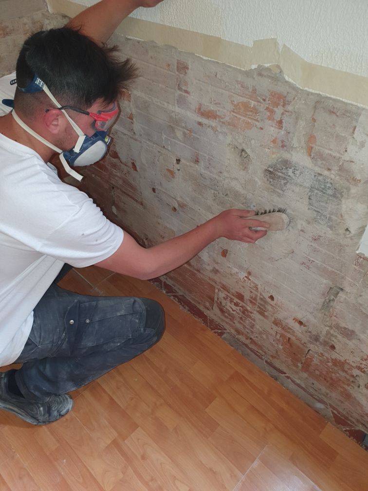 Un homme porte un masque de protection et des lunettes de sécurité pendant qu'il applique un produit sur un mur en briques apparentes.