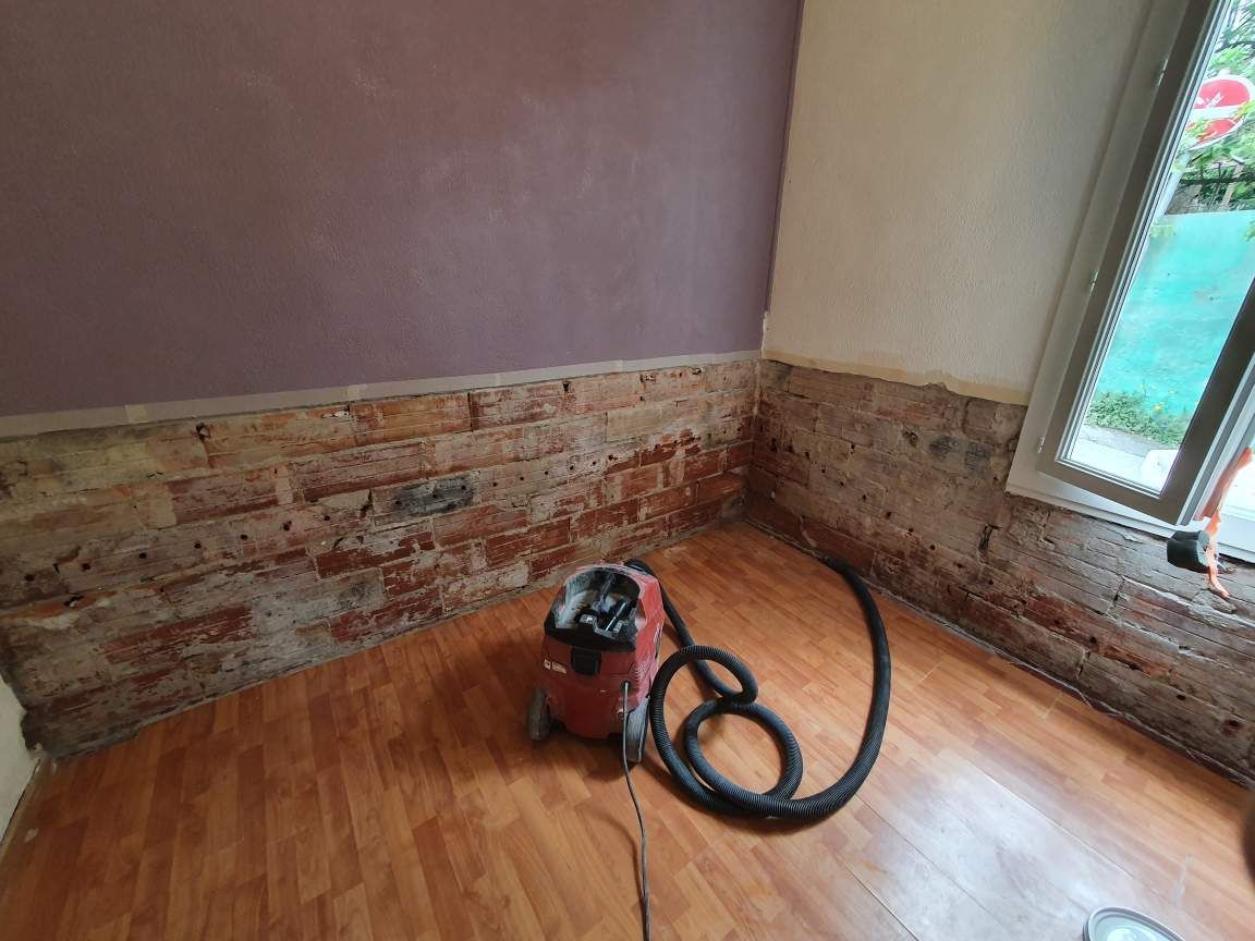 Un aspirateur industriel posé sur un sol en parquet dans une pièce en rénovation. Les murs montrent des briques apparentes et des zones de plâtre enlevé.
