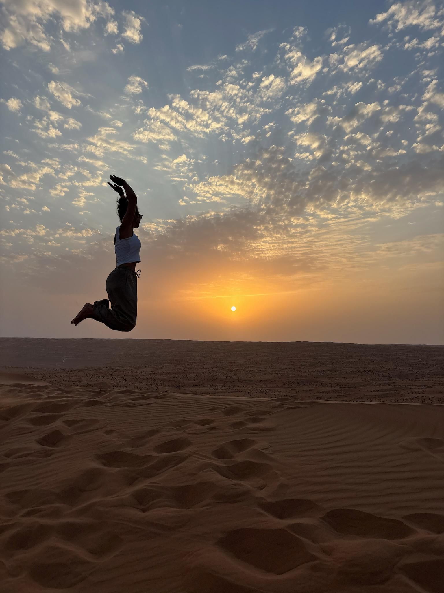 Eine Person springt als Silhouette an einem Sandstrand bei Sonnenuntergang unter einem wolkenverhangenen Himmel