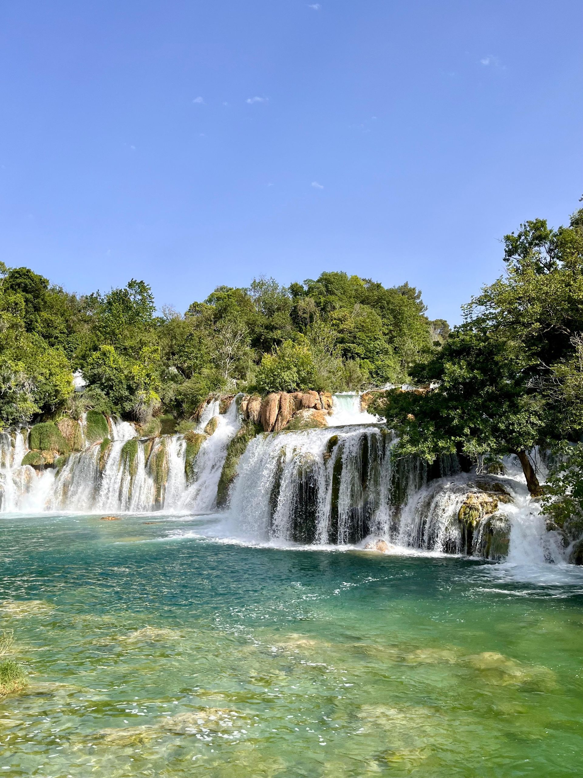 Ein gestufter Wasserfall mündet in ein klares, türkisfarbenes Becken unter einem strahlend blauen Himmel