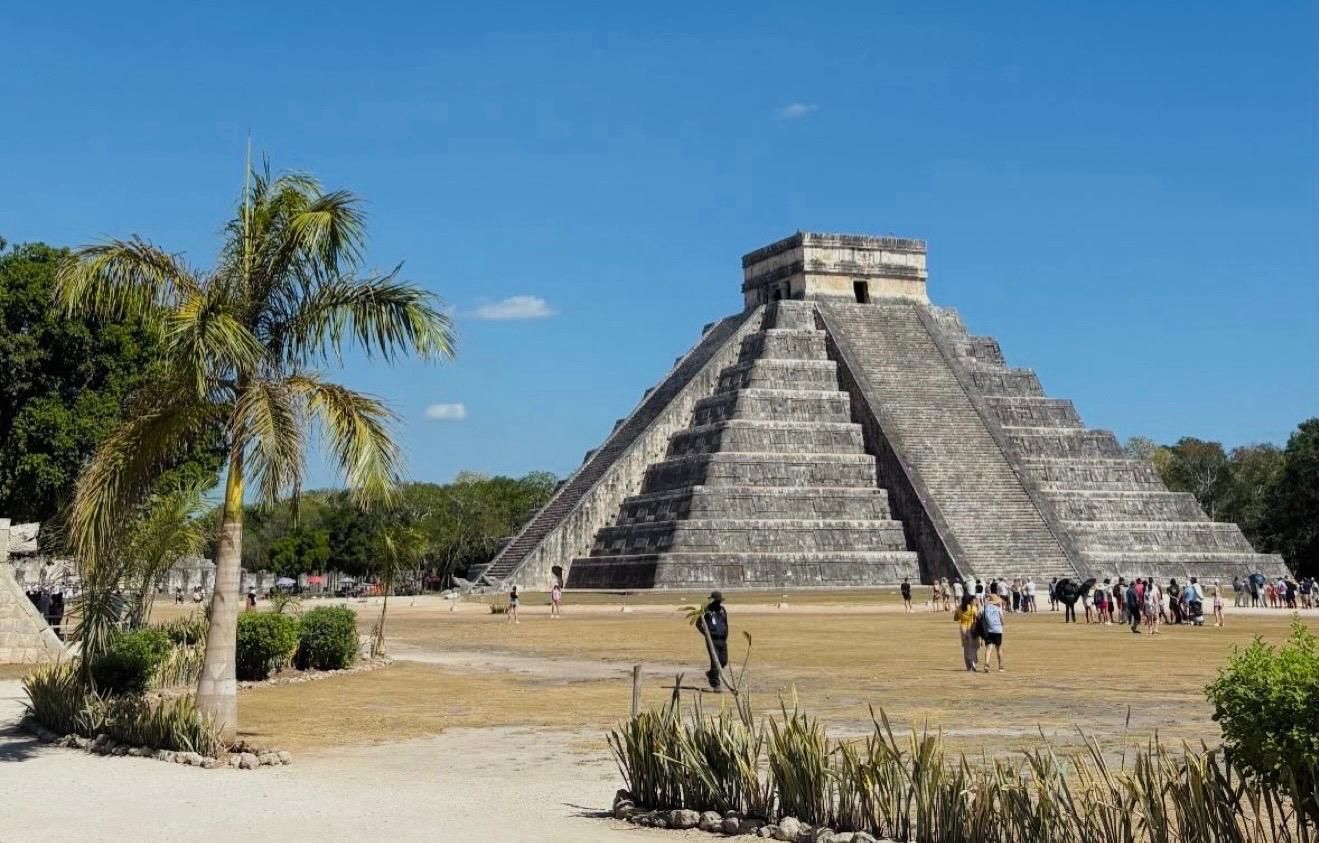 Maya-Pyramide in Chichén Itzá unter blauem Himmel, mit einer Palme und Besuchern im Vordergrund