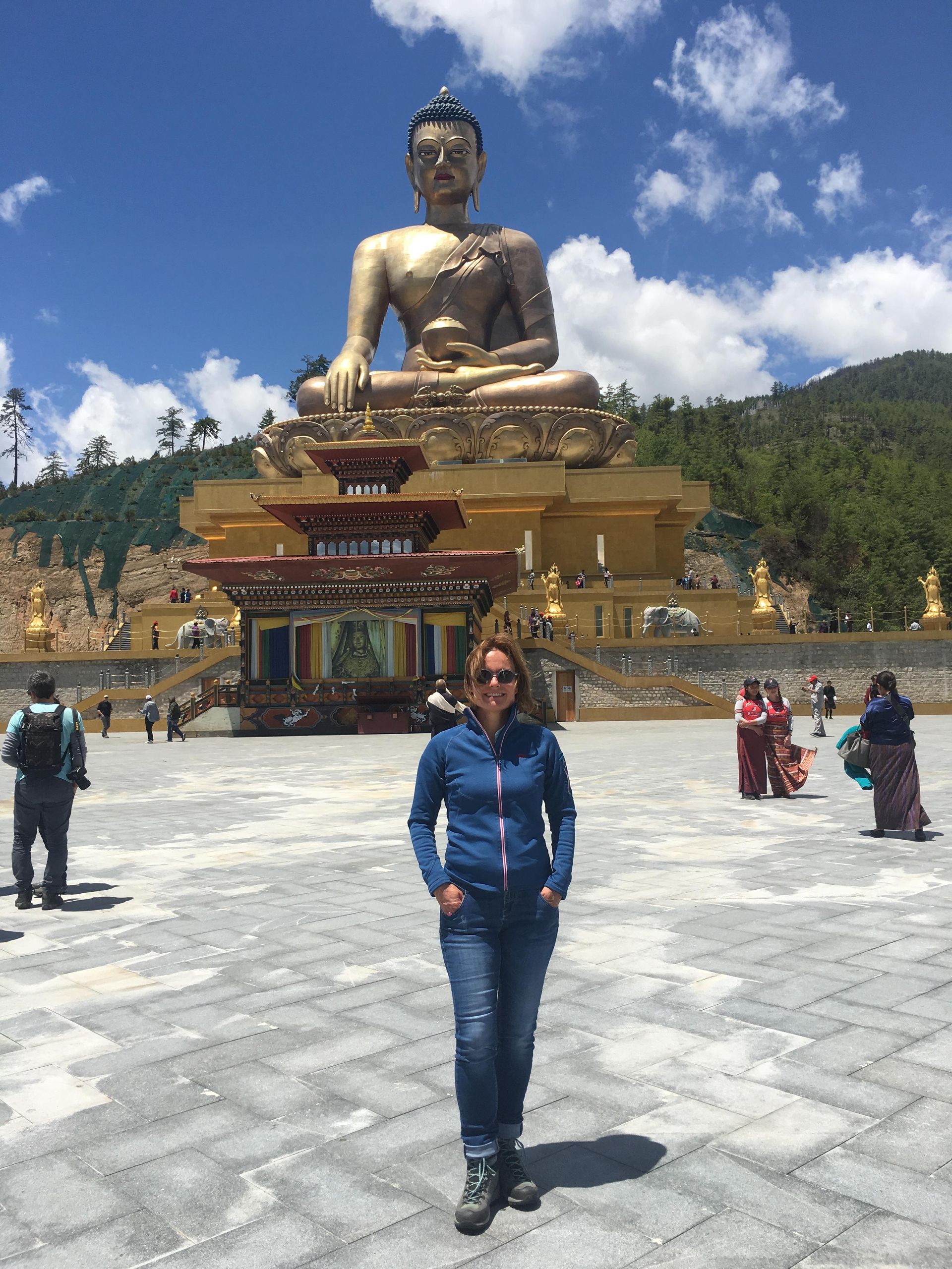 Eine Person steht vor einer großen goldenen Buddha-Statue in einem Bergtempel unter blauem Himmel.