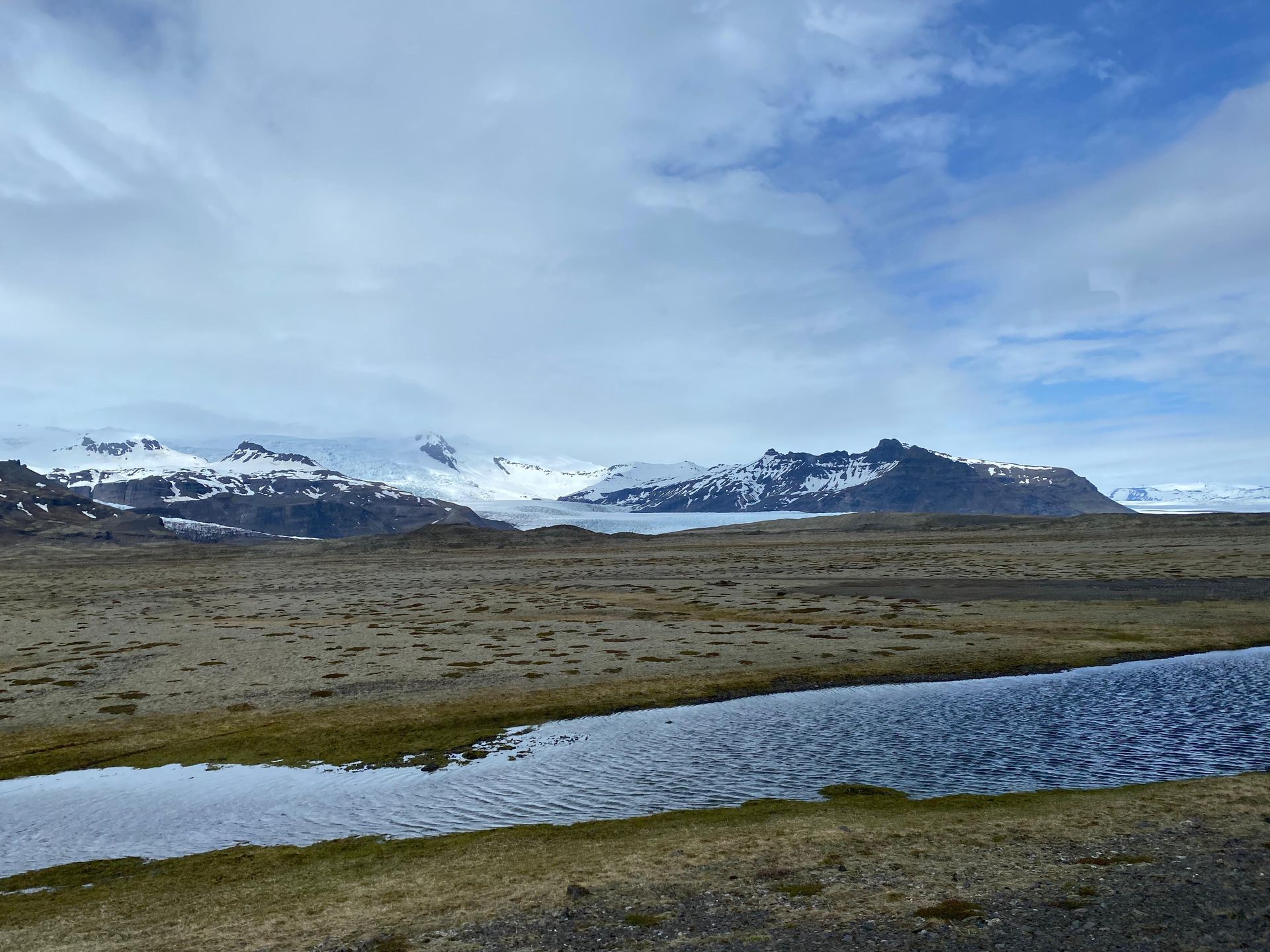 Schneebedeckte Berge hinter einer grasbewachsenen Ebene mit einem gewundenen Bach unter einem bewölkten blauen Himmel