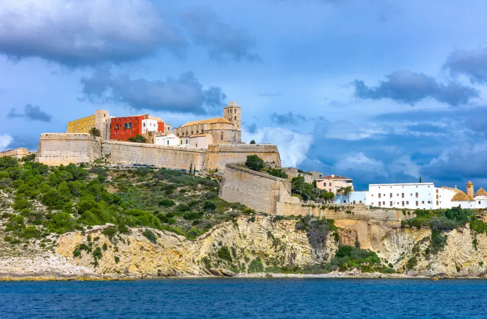 Fortaleza costera en la cima de una colina y edificios blancos sobre el mar bajo un cielo azul nublado.