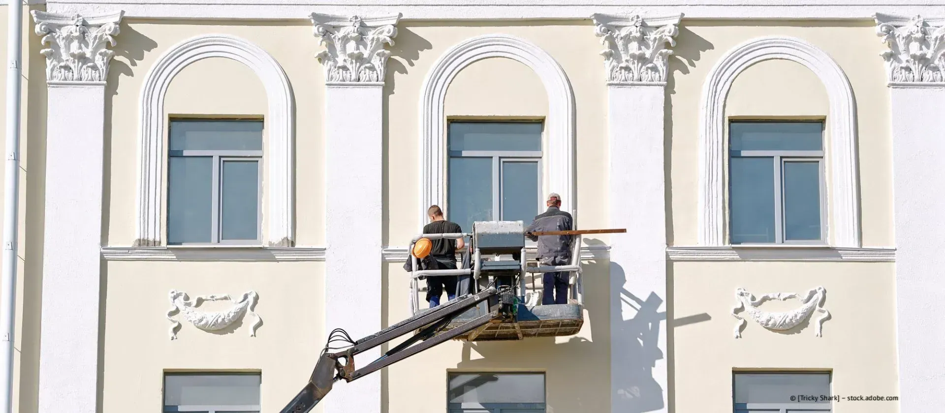 Zwei Arbeiter reparieren auf einer Hebebühne die reich verzierte weiße Gebäudefassade in der Nähe eines Fensters.