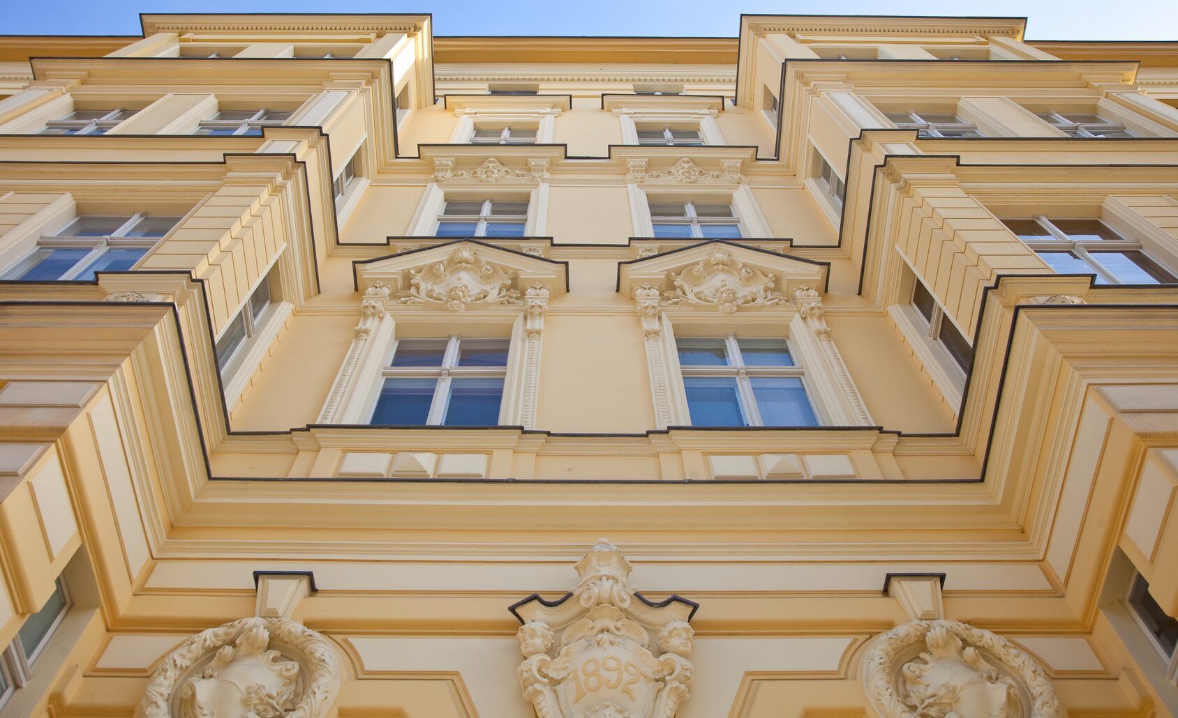Blick nach oben auf eine beige, reich verzierte Gebäudefassade mit Fenstern und Stuckdetails