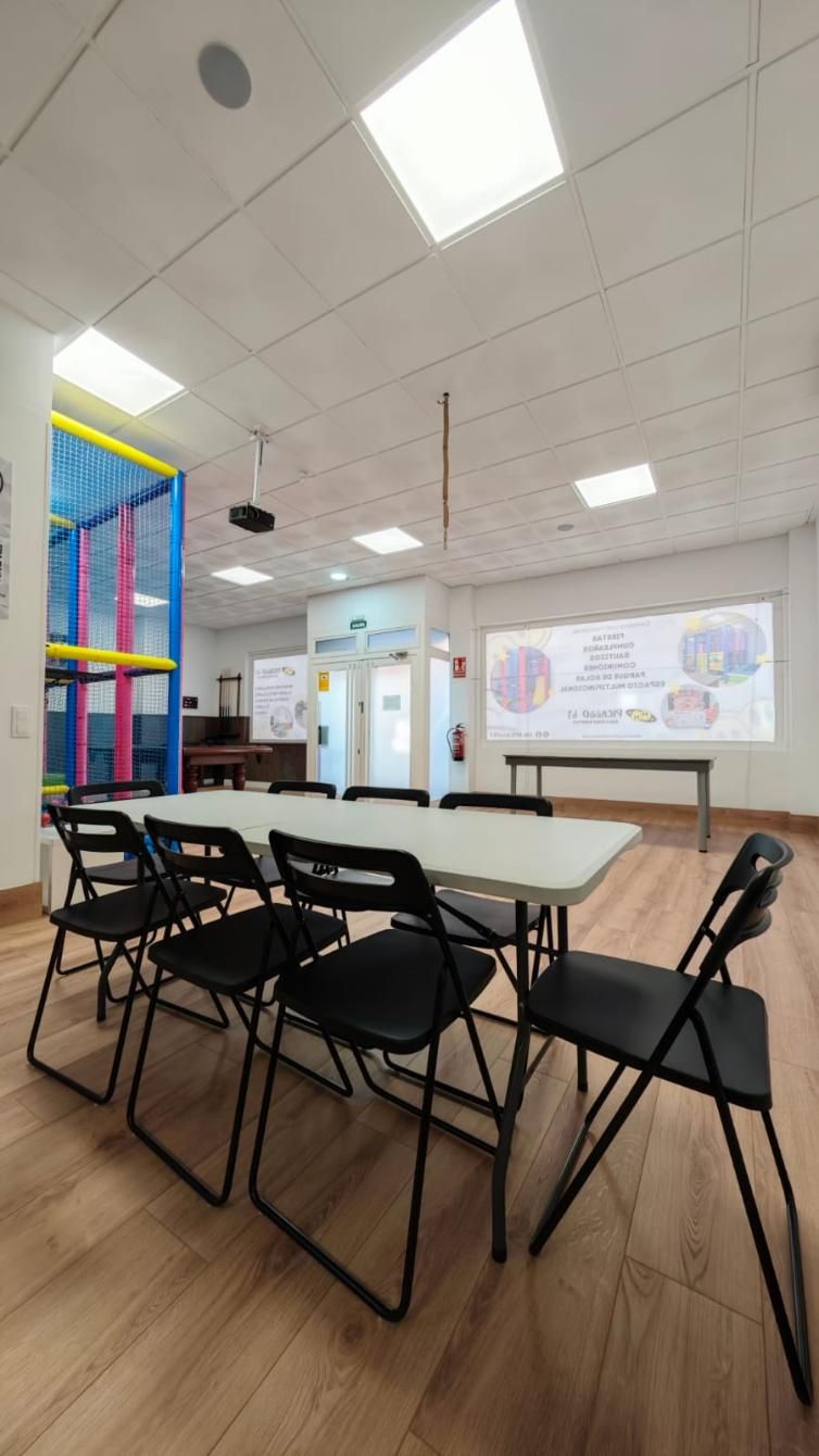 Un aula con una mesa y sillas, una pantalla de proyección y una estructura de juegos visible al fondo.