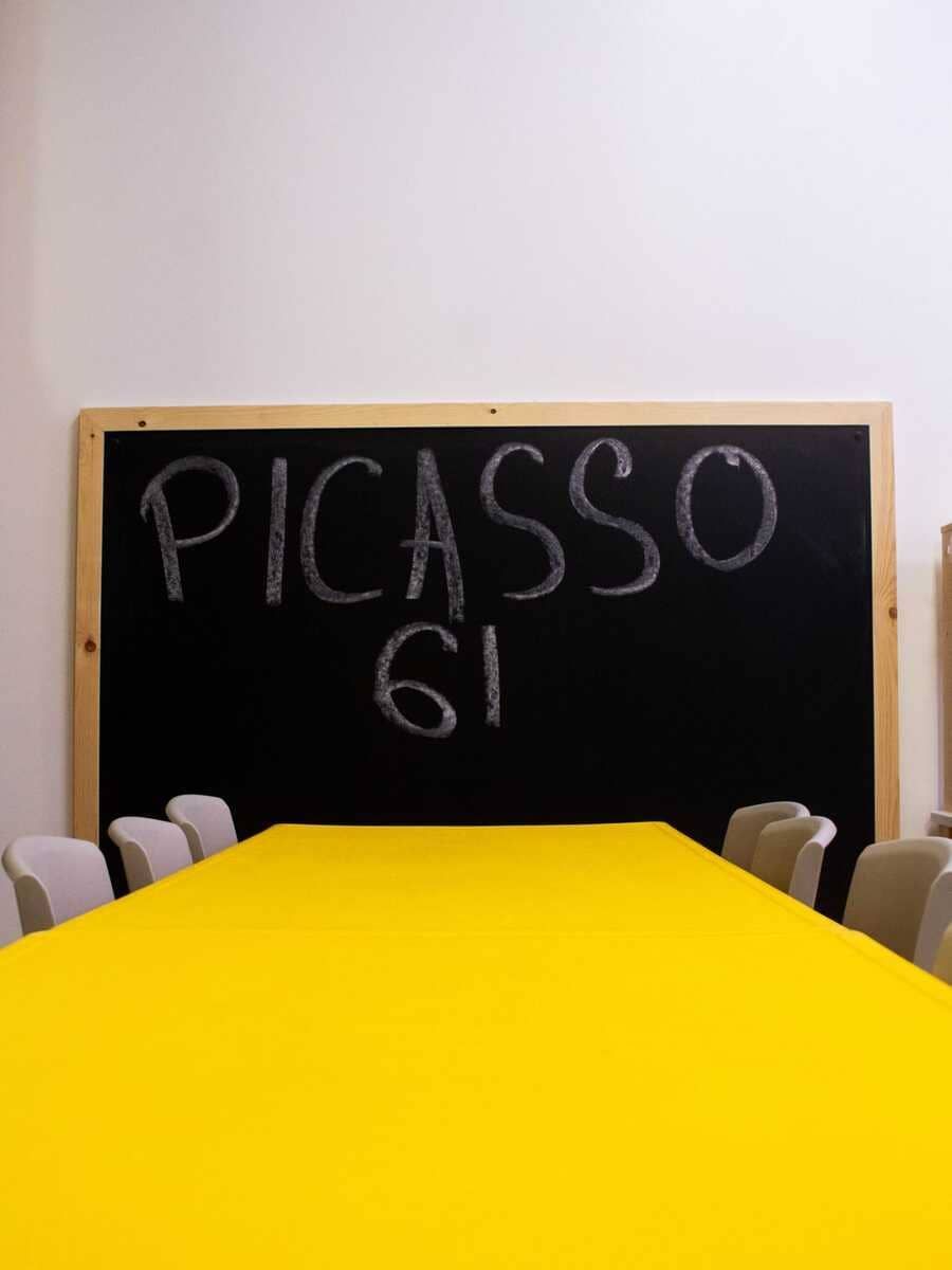 Pizarra con la inscripción “PICASSO 61” detrás de una mesa de conferencias de color amarillo brillante con sillas a ambos lados.