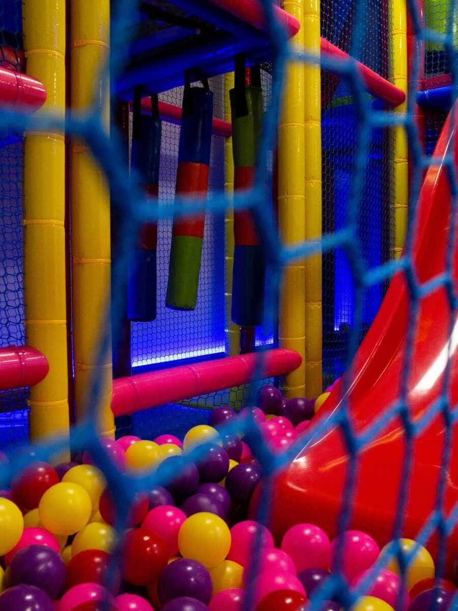 Zona de juegos interior colorida con red, postes acolchados y una piscina de bolas con pelotas rojas, amarillas y rosas.
