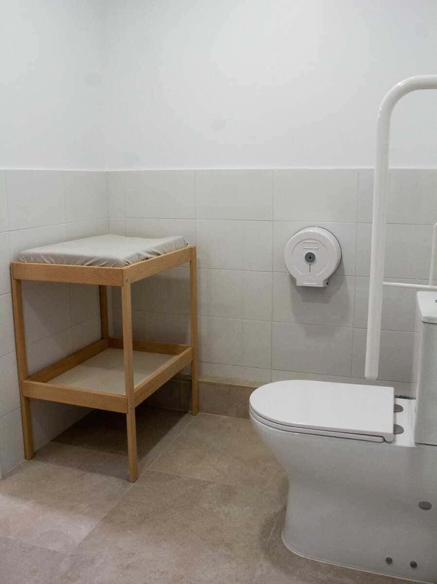 Baño accesible con inodoro suspendido, barras de apoyo y un pequeño banco de madera.