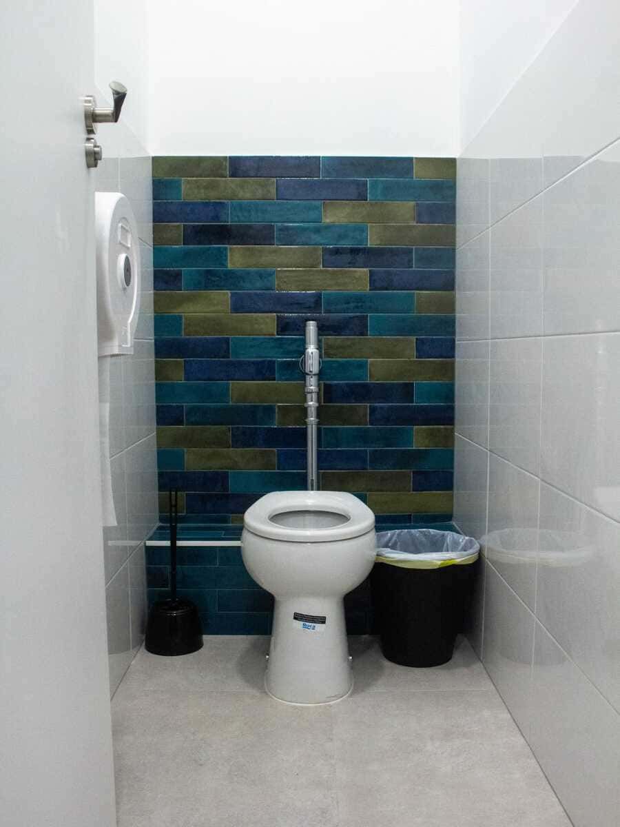 Baño pequeño con inodoro blanco, pared decorativa de azulejos azul verdoso y papeleras a ambos lados.