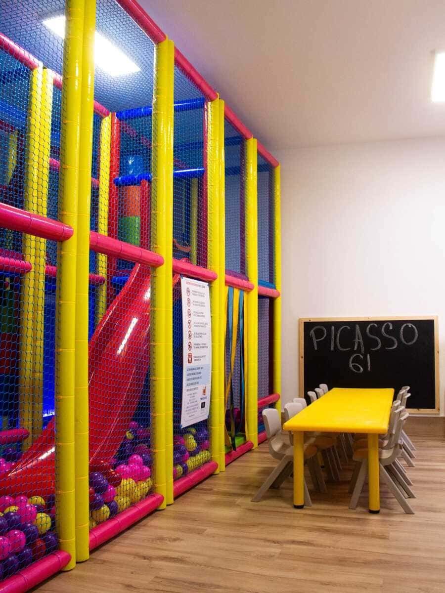 Zona de juegos interior colorida con estructuras para trepar con redes de color rosa, amarillo y azul, y una pequeña mesa cerca de un cartel de pizarra.