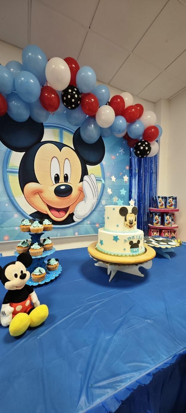 Decoraciones para fiesta de cumpleaños con temática de Mickey Mouse: pastel, cupcakes, globos y peluche sobre un mantel azul.