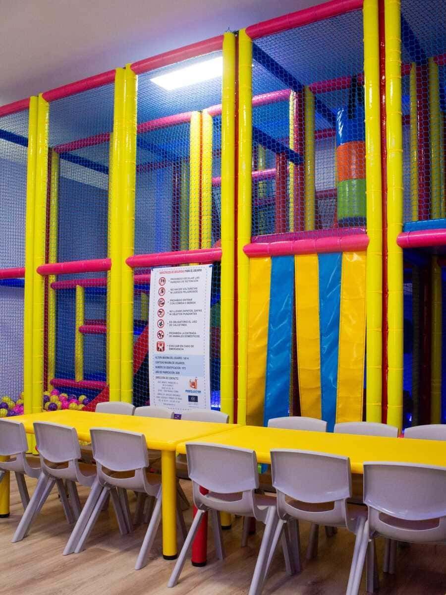 Zona de juegos interior para niños con estructuras acolchadas para trepar de color amarillo brillante, rosa y azul, y mesas en la parte delantera.