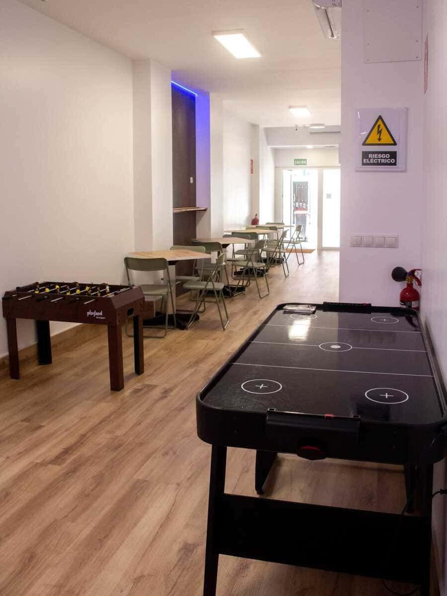 Mesas de air hockey en un pasillo luminoso con sillas a lo largo de la pared y un cartel de advertencia cerca de la puerta.