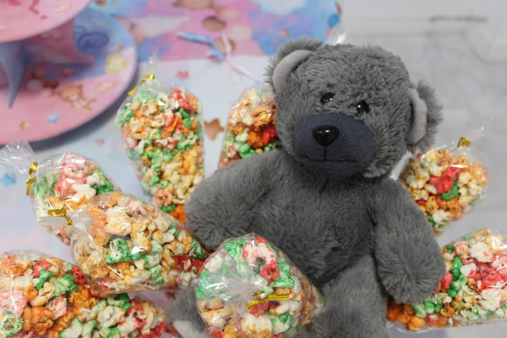 Osito de peluche gris rodeado de palomitas de maíz de colores en bolsas transparentes en una fiesta.