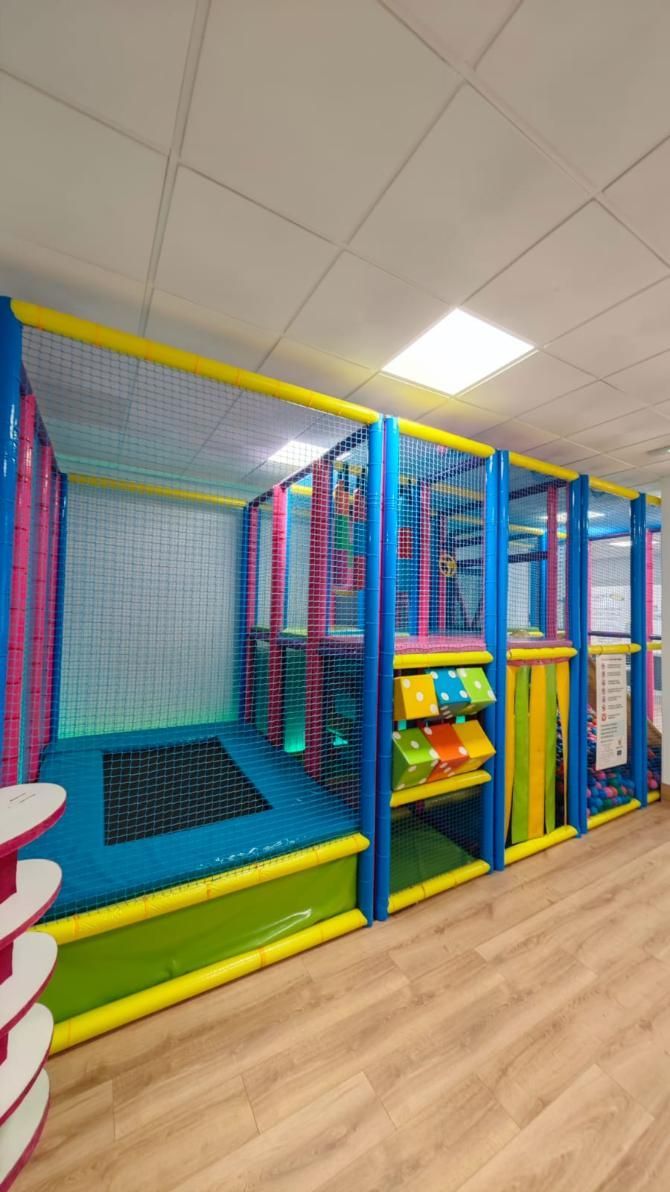 Parque infantil interior con estructuras acolchadas de color azul, amarillo y verde, redes y un pequeño trampolín.