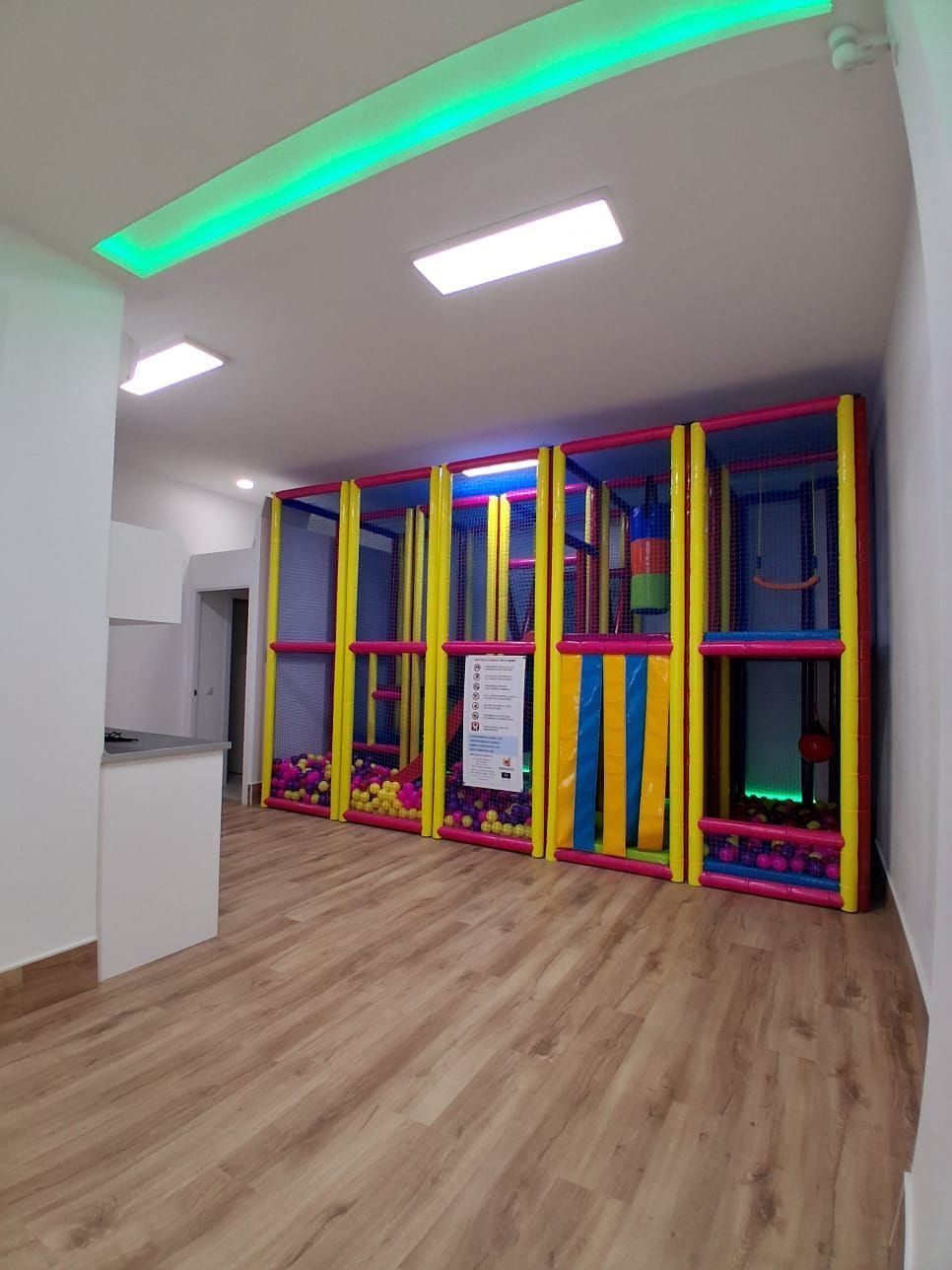 Zona de juegos interior luminosa con colorida estructura acolchada para trepar y suelo de madera bajo luces de techo verdes.