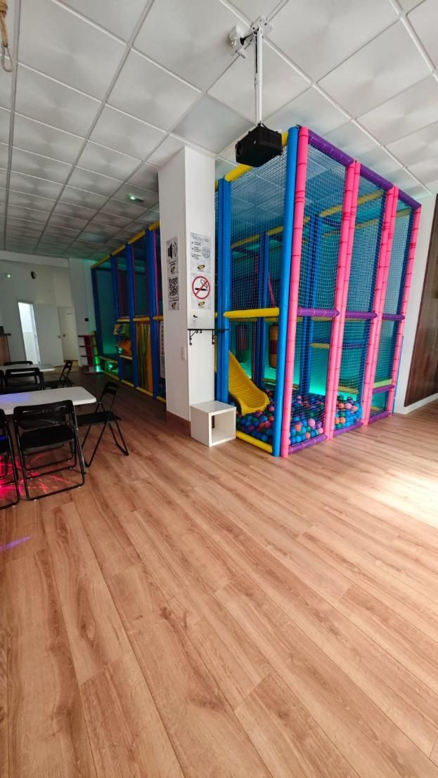 Zona de juegos infantiles interior con coloridas estructuras para escalar, tobogán y suelo de madera.
