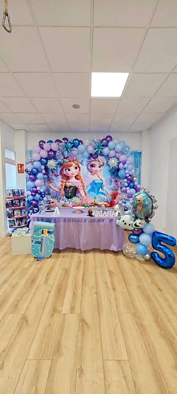 Fiesta de cumpleaños con temática de Frozen: Fondo con Elsa y Anna, mesa con golosinas, globos y globos con el número 5.