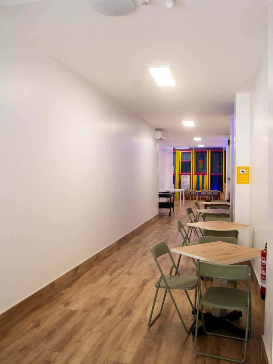 Largo pasillo con mesas y sillas a lo largo de la pared derecha, paredes blancas, suelo de madera y una colorida ventana al final.
