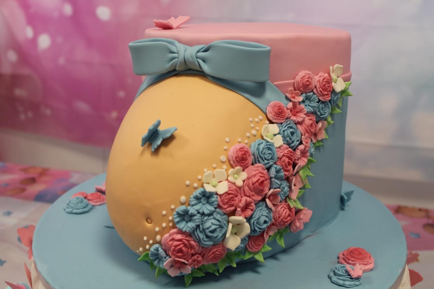 Pastel decorado en forma de barriga de embarazada con lazo azul, flores rosas y azules y mariposas.