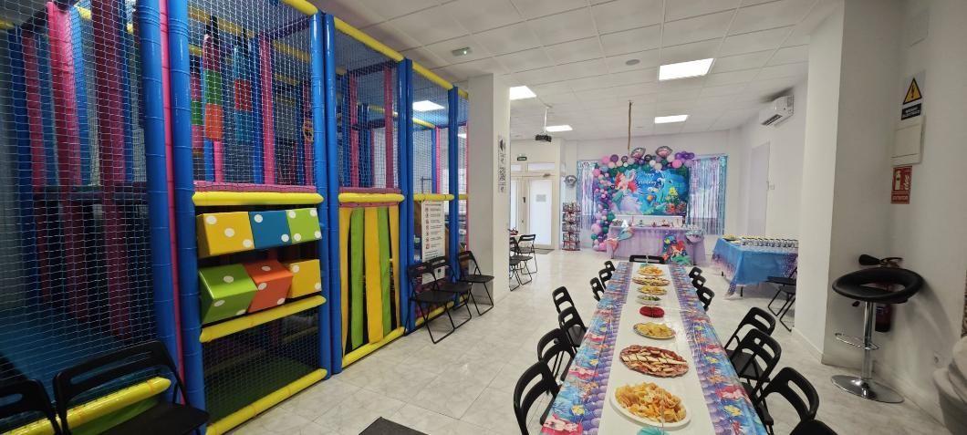 Parque infantil interior con decoraciones de fiesta, mesa larga con comida y sillas y una estructura de juegos.