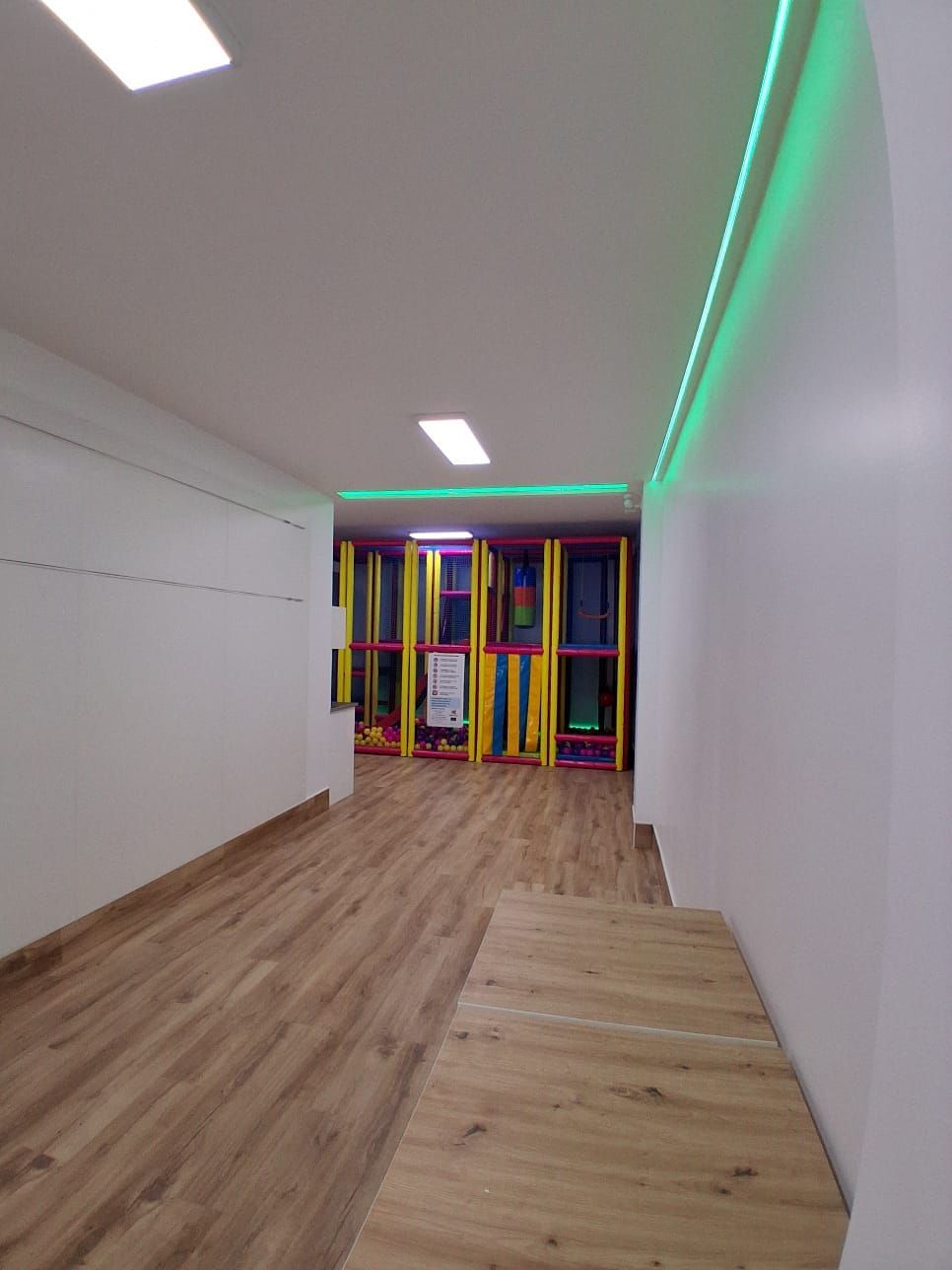Pasillo interior vacío con suelo de madera, paredes blancas y una colorida zona de juegos al final.