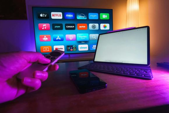 Persona que sostiene los controles remotos de un televisor, una tableta y un teléfono sobre un escritorio, todos iluminados con luz violeta.
