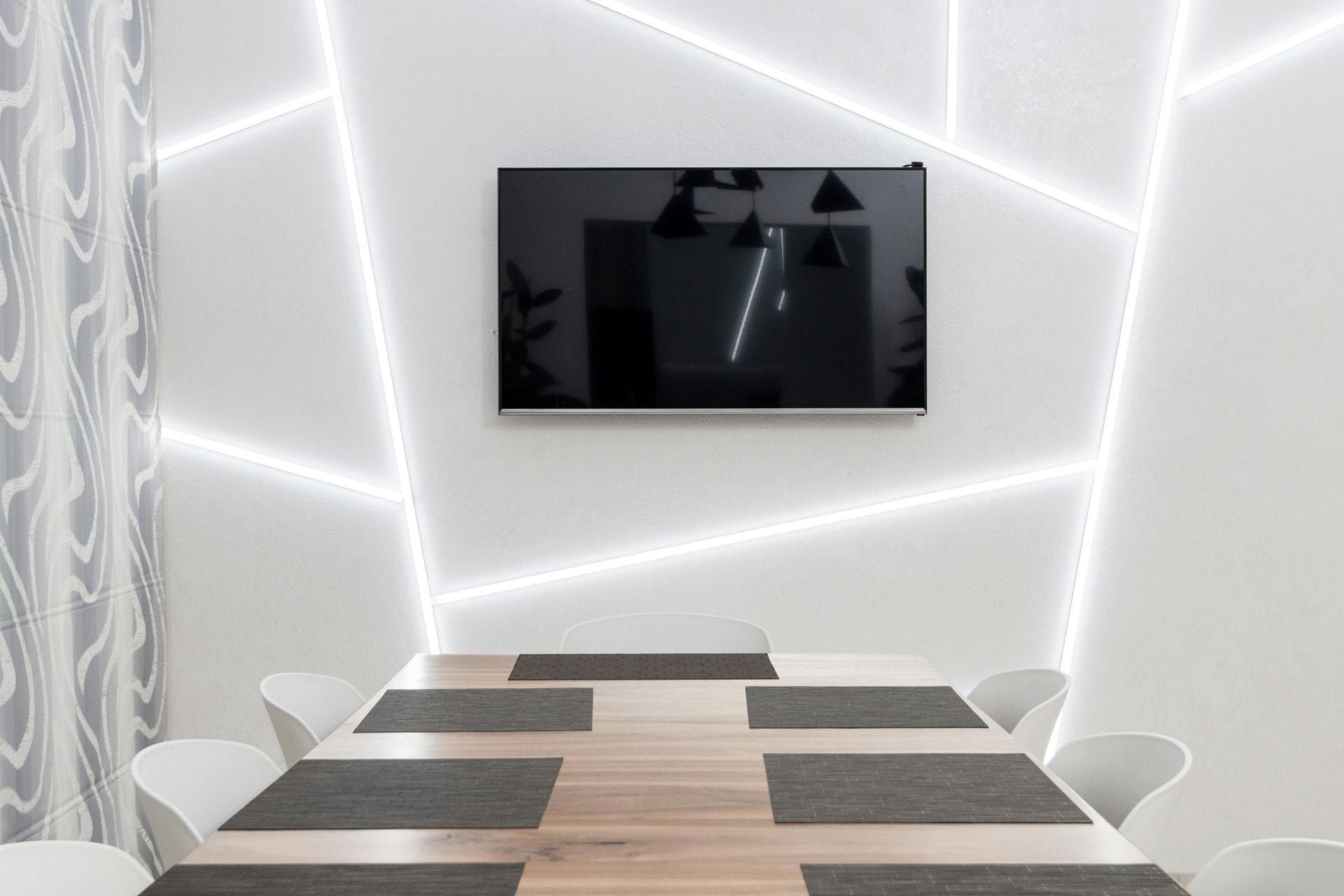 Comedor con TV montada en pared blanca con iluminación LED, encima de una mesa de madera y sillas blancas.