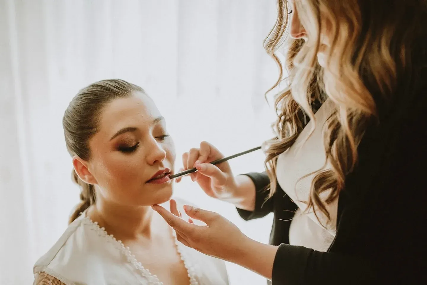 Maquilladora aplicando lápiz labial a una mujer, entorno interior con luz natural.