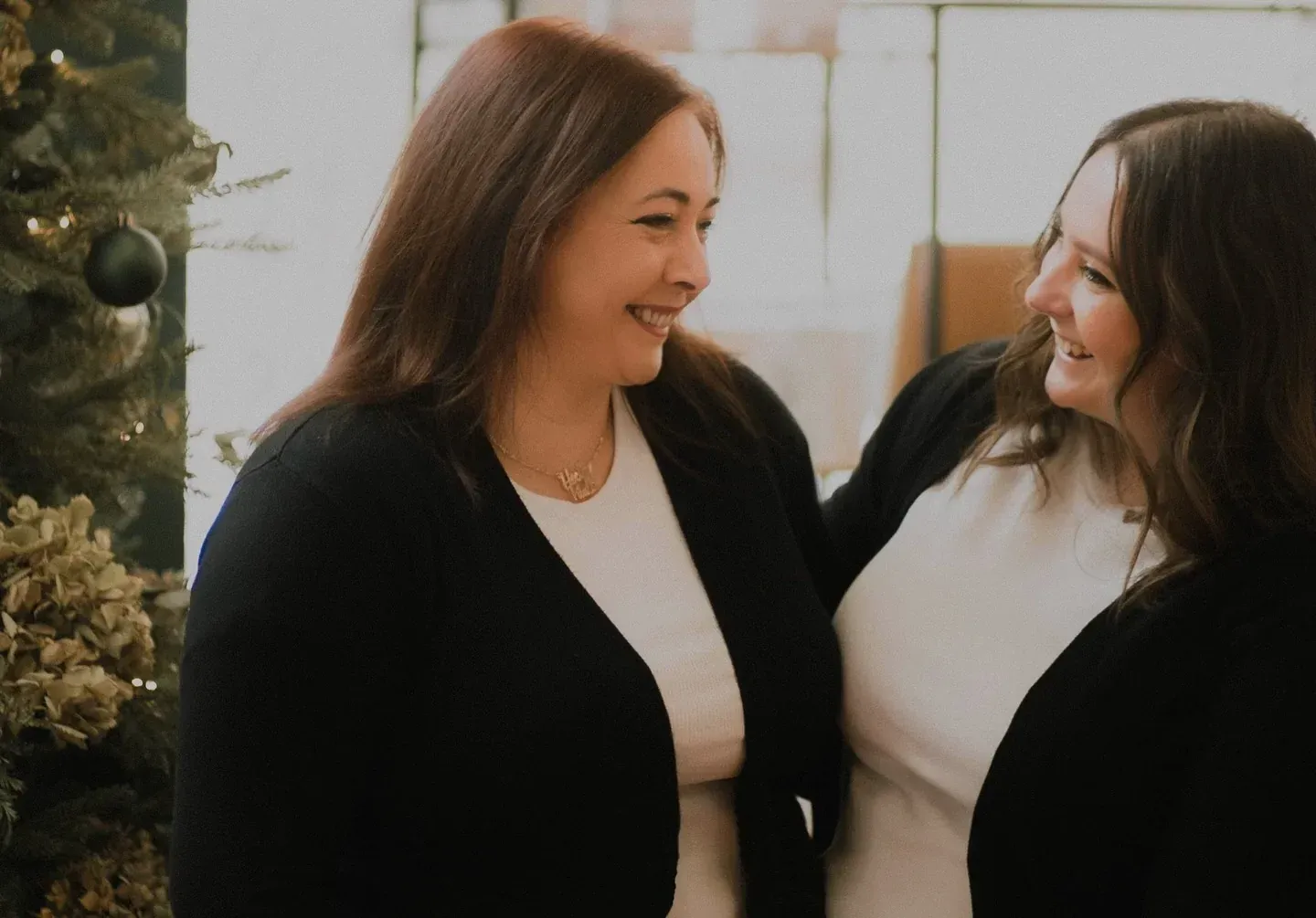 Dos mujeres sonriendo y abrazándose, cerca de un árbol de Navidad decorado.