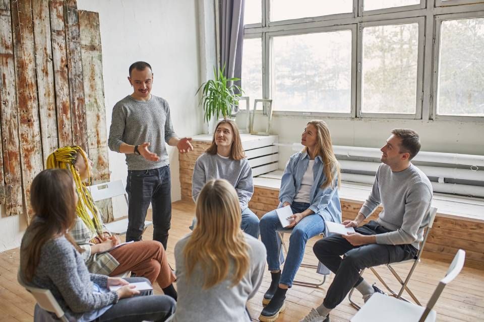 Bijeenkomst in een kleine groep in een lichte ruimte, met een spreker die staat en de anderen die in een kring zitten.