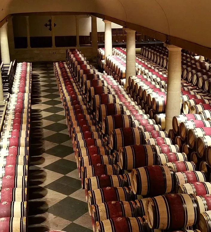 Filas de barriles de vino en una bodega con suelo a cuadros, columnas y un emblema en la pared.