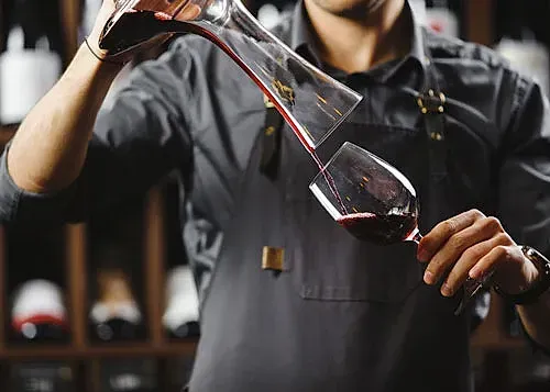 Persona con delantal vertiendo vino tinto de un decantador en una copa, con un botellero al fondo.