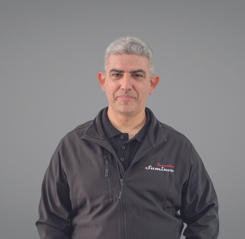 Un hombre que lleva una chaqueta negra y un polo negro está parado frente a una pared gris.