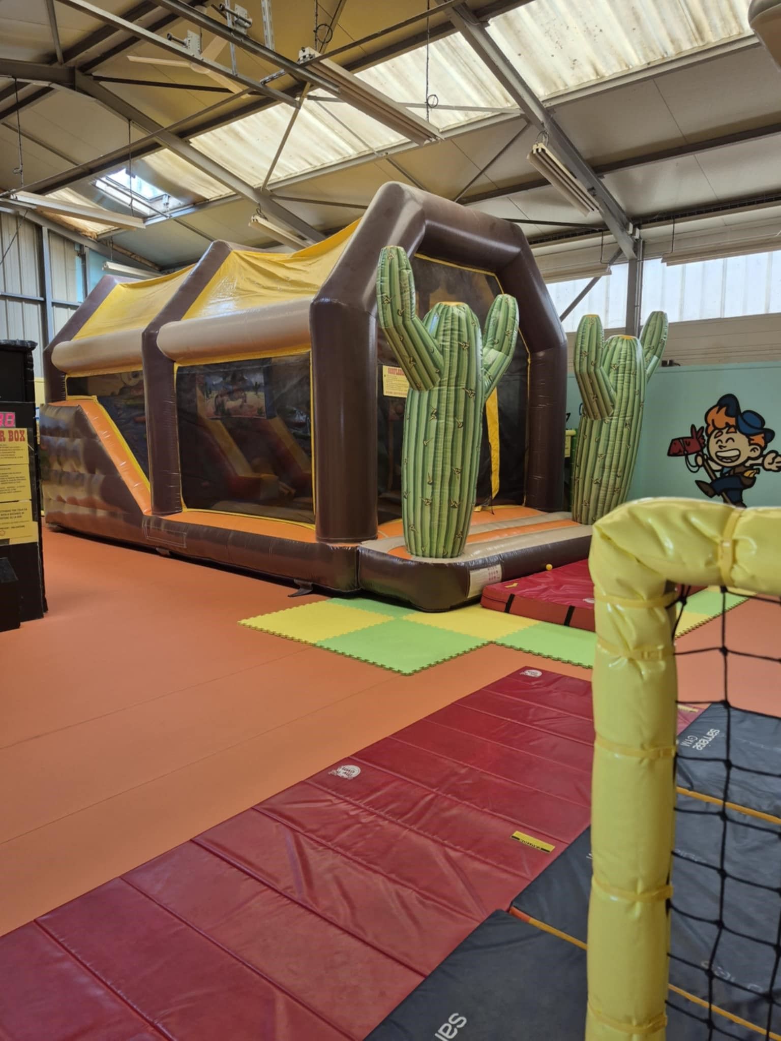 Une aire de jeux intérieure avec une structure gonflable marron sur le thème du Far West ; toboggan et détails de cactus.