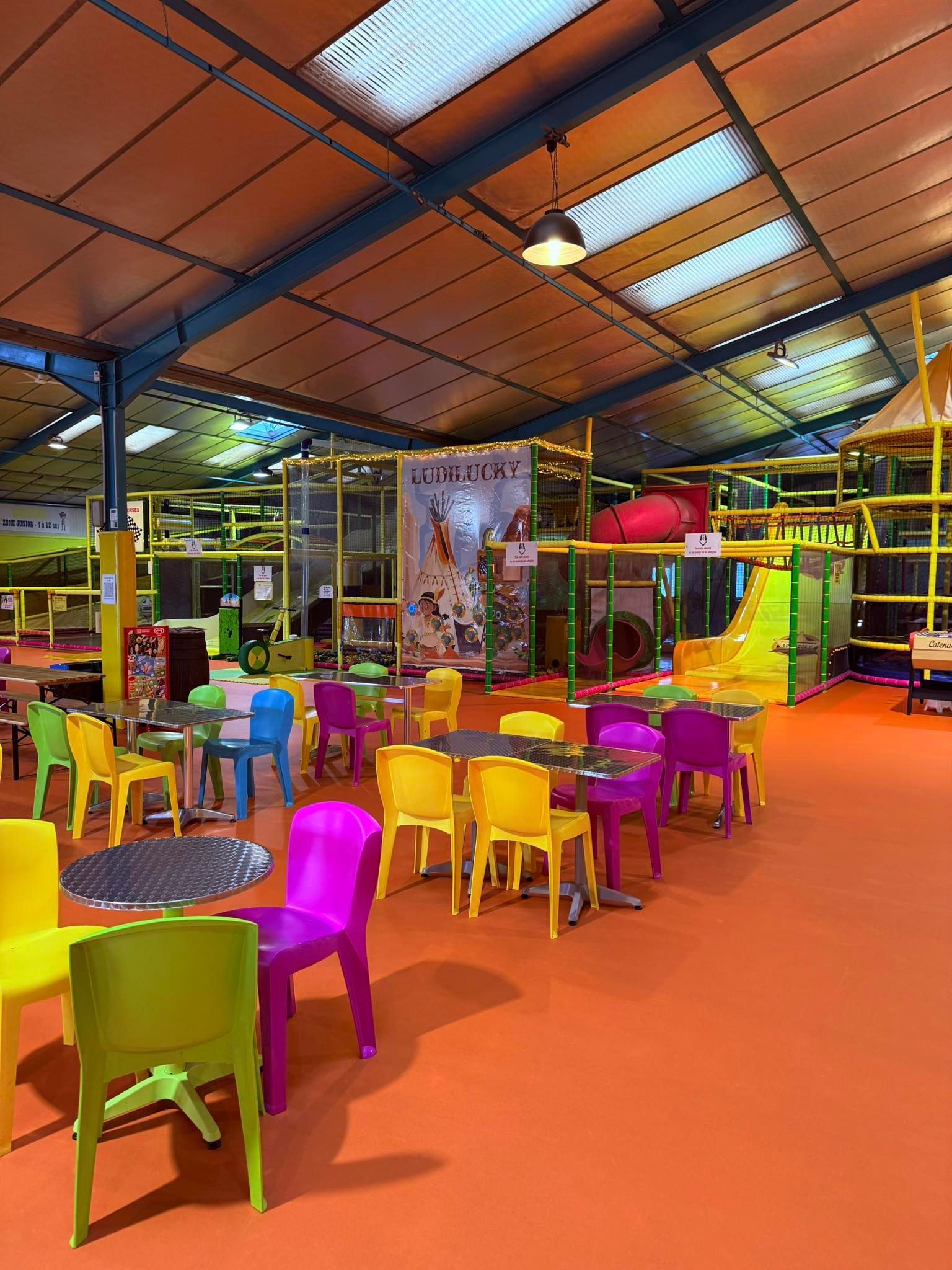 Aire de jeux intérieure avec chaises colorées et structures de jeux ; sol orange et haut plafond.