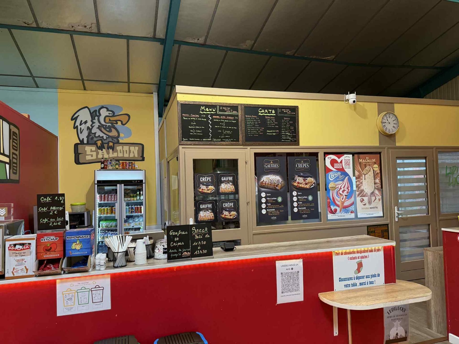 Comptoir de restaurant avec murs jaunes, bordures rouges, menus affichés et réfrigérateur rempli de boissons.