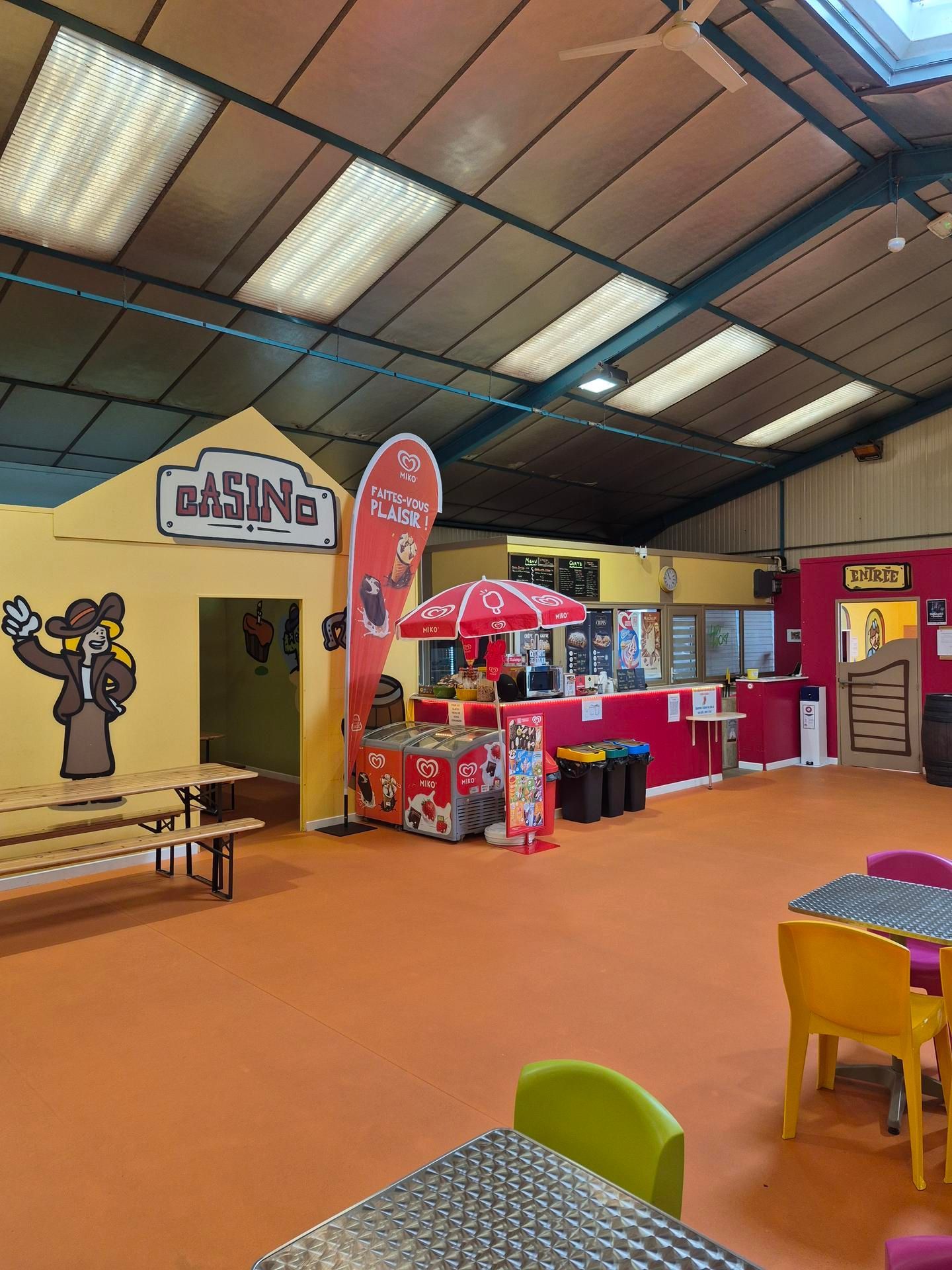 Aire de jeux intérieure avec tables colorées et kiosques alimentaires sous un haut plafond.