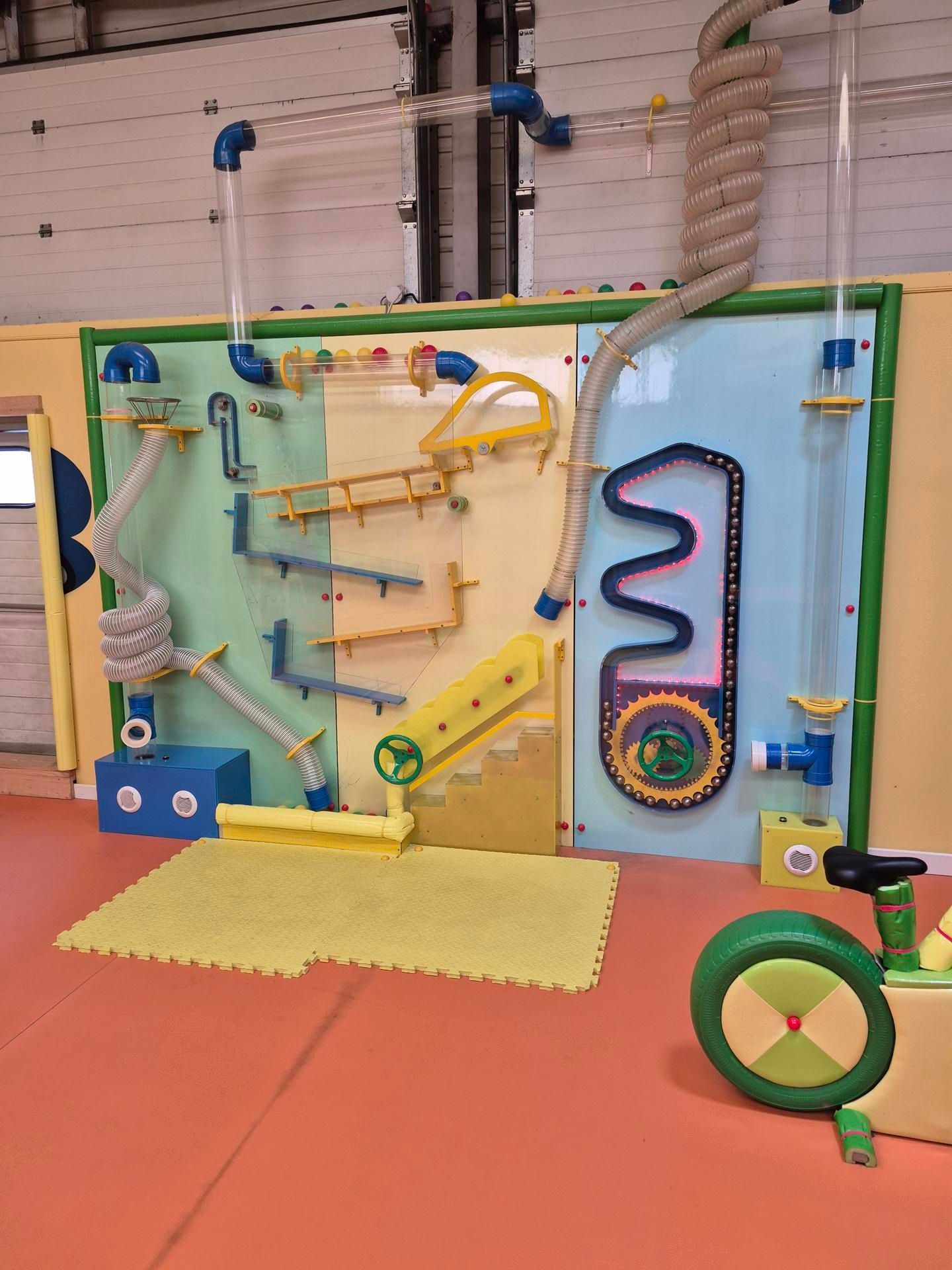 Mur de jeux avec tuyaux, rampes et vélo. Multicolore, fixé sur un mur.