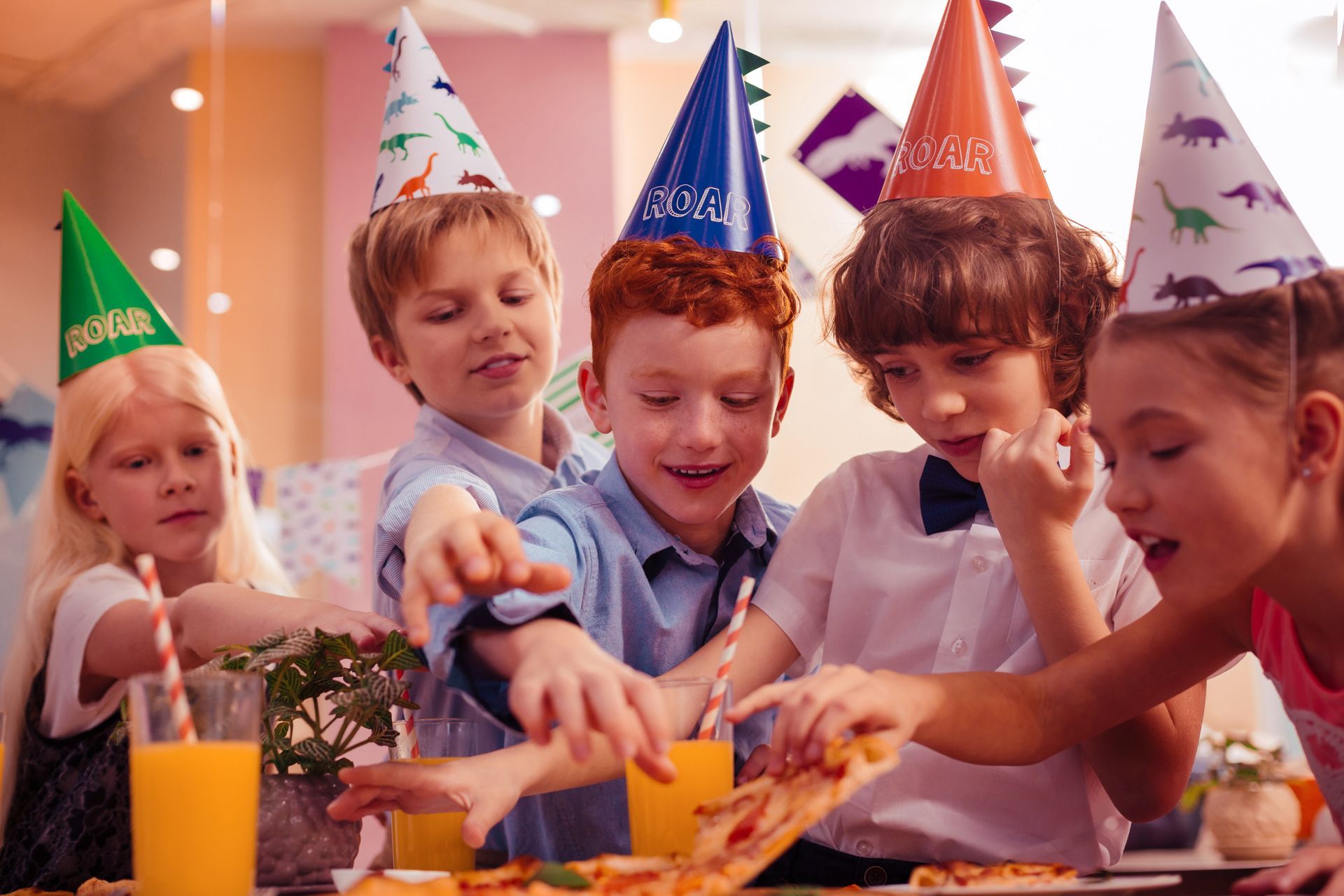 Des enfants coiffés de chapeaux de fête tendent la main vers des parts de pizza lors d'une fête, du jus d'orange est posé sur la table.